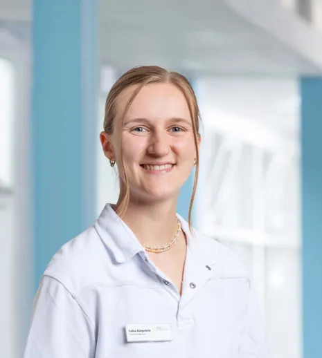 Porträt einer lächelnden Frau in weißem Arztkittel mit Namensschild vor hellem, blauem Klinikflur.