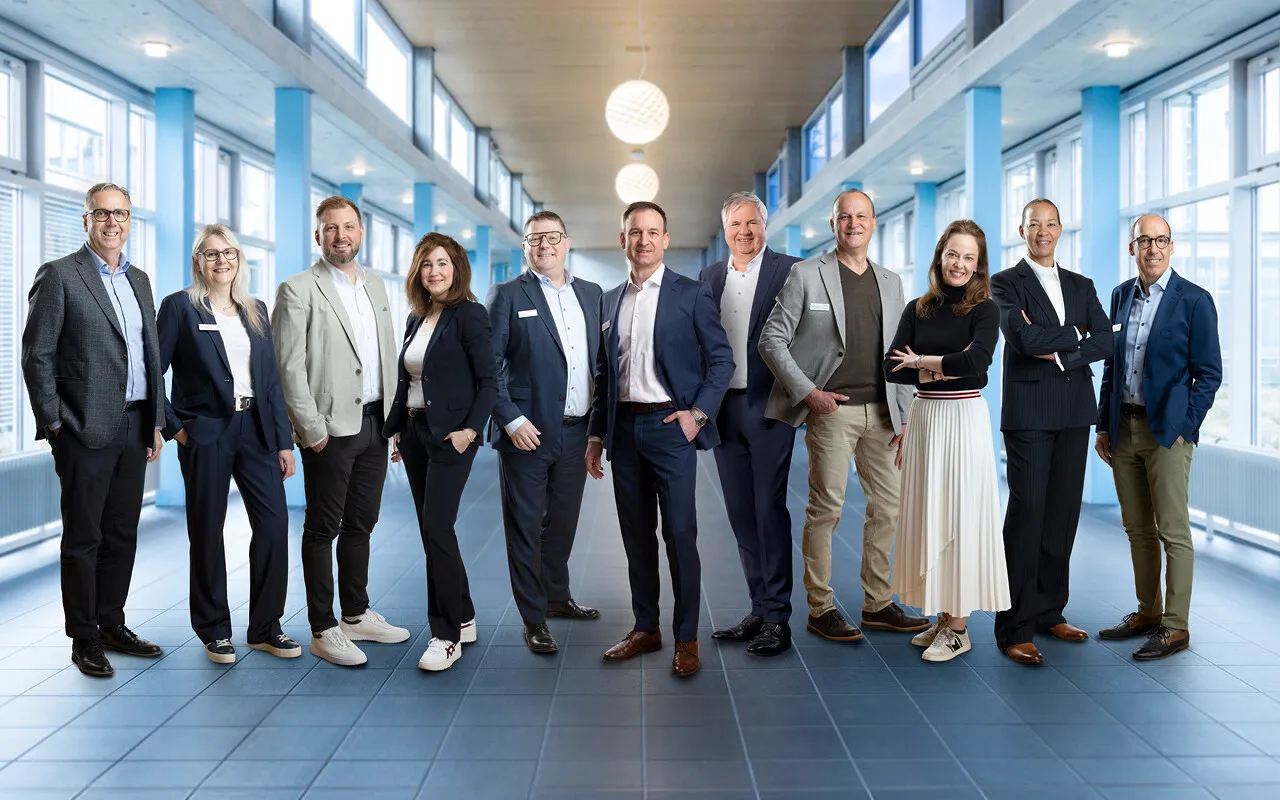 Gruppenfoto mehrerer Personen in Business-Kleidung in einem hellen Büroflur mit großen Fenstern und blau akzentuierten Wänden.