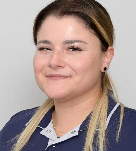 Porträt einer lächelnden Frau mit braun-blondem Haar, schwarzem Ohrstecker und dunkelblauer Arbeitskleidung vor grauem Hintergrund.