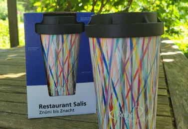 Bunte Mehrwegkaffeetasse mit schwarzem Deckel, neben einer blauen Verpackung mit dem Schriftzug 