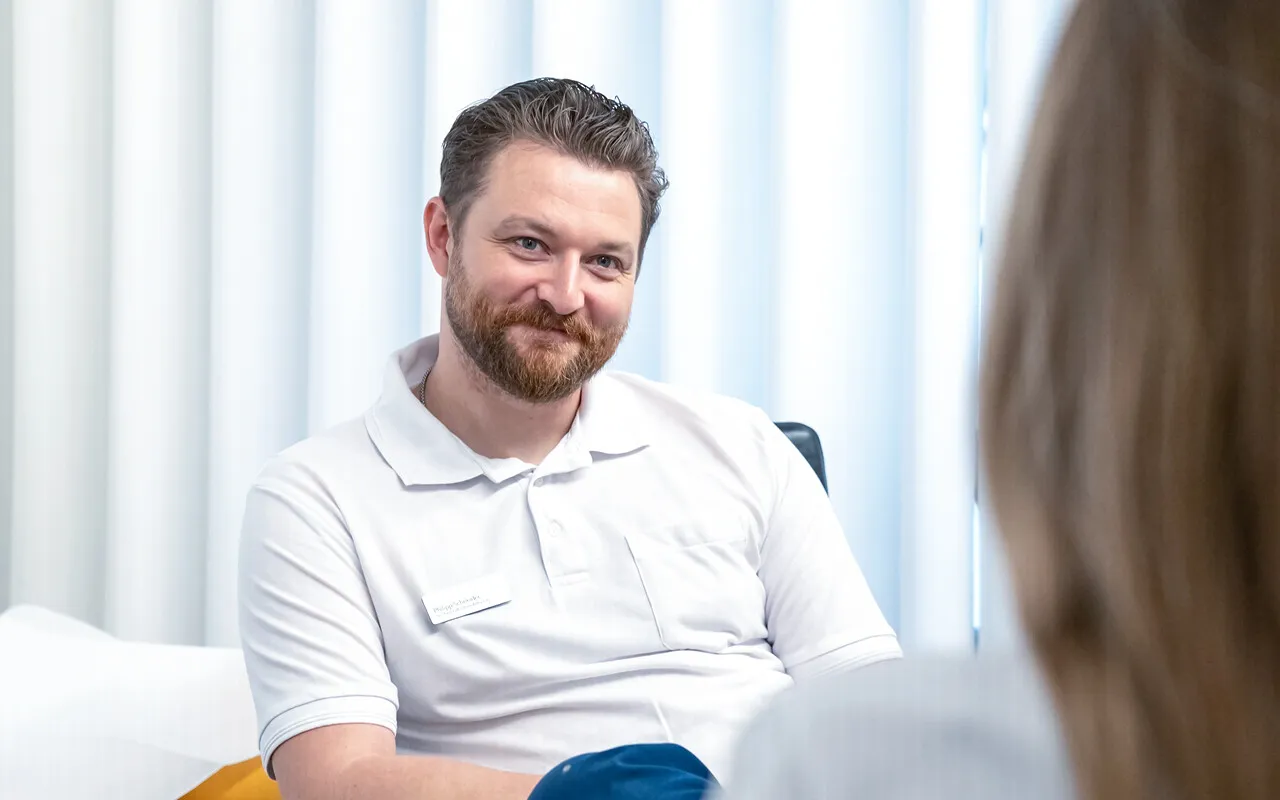 Männlicher Arzt im weißen Poloshirt mit Namensschild sitzt im Gespräch im Behandlungsraum, freundliches Lächeln; im Hintergrund helle Vorhänge und eine weitere Person im Vordergrund.