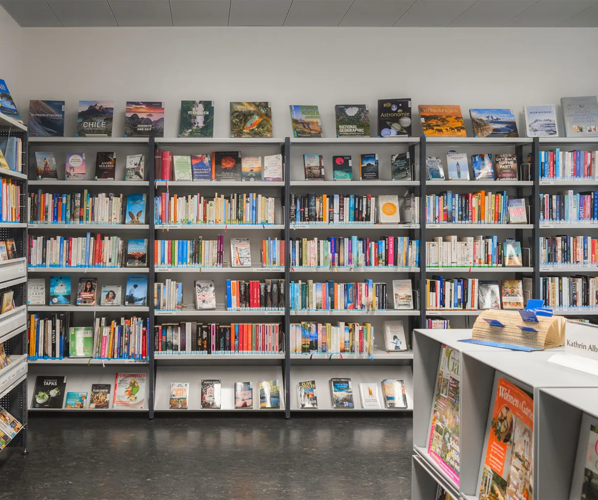 Regal mit einer Vielzahl von Büchern in unterschiedlichen Farben und Größen, darunter Sachbücher und Romane. Ein Tisch mit einer Namenskarte und einem kleinen Hut steht im Vordergrund, während im Hintergrund weitere Bücherregale sichtbar sind. Die Wänd...