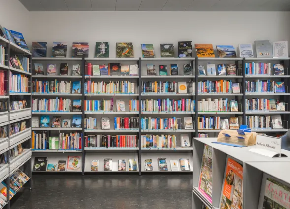 Regal mit einer Vielzahl von Büchern in unterschiedlichen Farben und Größen, darunter Sachbücher und Romane. Ein Tisch mit einer Namenskarte und einem kleinen Hut steht im Vordergrund, während im Hintergrund weitere Bücherregale sichtbar sind. Die Wänd...