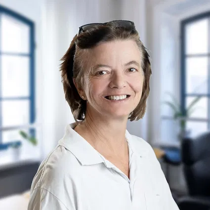 Lächelnde Frau mit kurzen braunen Haaren und Brille auf dem Kopf, in weißem Hemd vor hellen Bürofenstern.