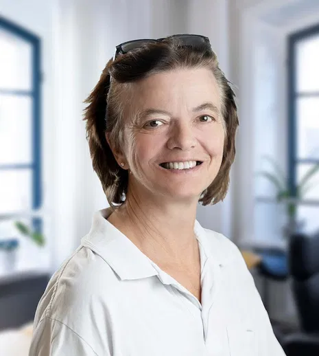 Lächelnde Frau mit kurzen braunen Haaren und Brille auf dem Kopf, in weißem Hemd vor hellen Bürofenstern.