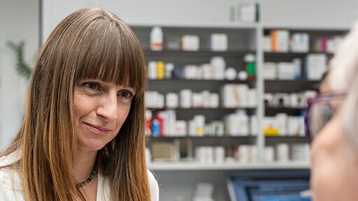 Frau mit braunem Haar und Pony im Porträt vor einer Regalschrankwand mit Büchern und Medikamenten.
