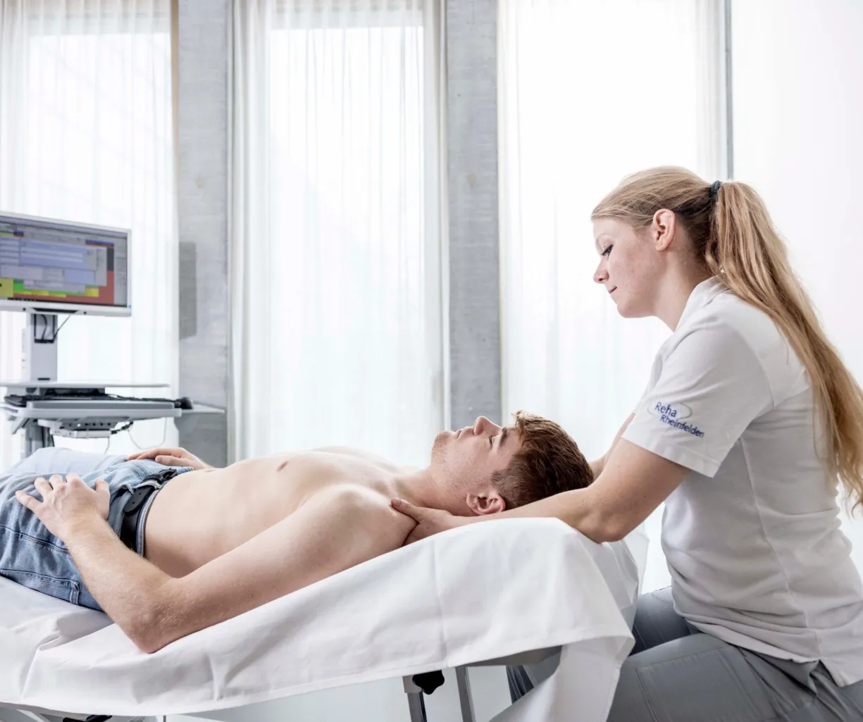 Mann liegt auf einer Behandlungsliege, während eine Therapeutin ihm den Nacken massiert. Im Hintergrund steht ein Computer mit einem Bildschirm. Helle, moderne Umgebung mit großen Fenstern.