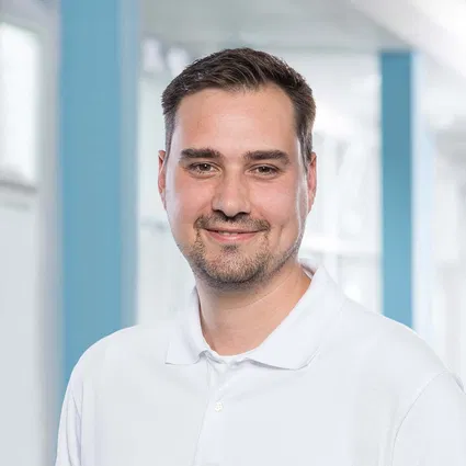 Porträt eines lächelnden Mannes mit kurzen dunklen Haaren und weißem Poloshirt vor hellem Hintergrund.