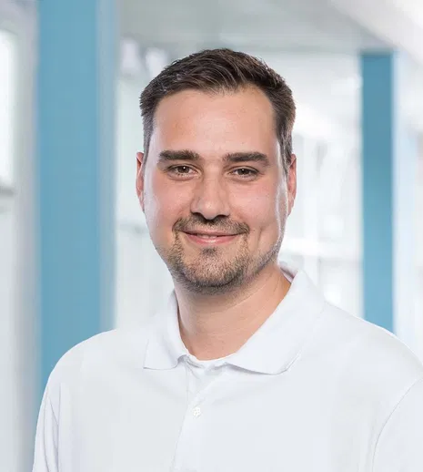 Porträt eines lächelnden Mannes mit kurzen dunklen Haaren und weißem Poloshirt vor hellem Hintergrund.