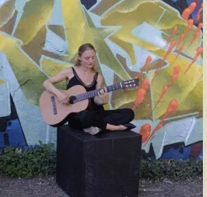 Frau sitzt barfuß auf einer schwarzen Kiste und spielt Gitarre. Im Hintergrund ist eine farbenfrohe Graffiti-Wand mit abstrakten Formen und Pflanzen zu sehen.