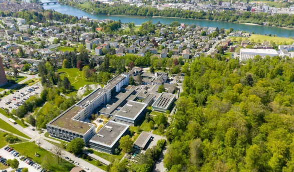 Luftaufnahme eines grünen Klinik- bzw. Universitätsgeländes mit mehreren Gebäudekomplexen, Innenhöfen und Zufahrtsstraßen, umgeben von Wohngebieten und Flusslauf.