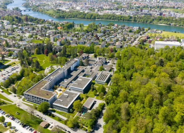 Luftaufnahme eines grünen Klinik- bzw. Universitätsgeländes mit mehreren Gebäudekomplexen, Innenhöfen und Zufahrtsstraßen, umgeben von Wohngebieten und Flusslauf.
