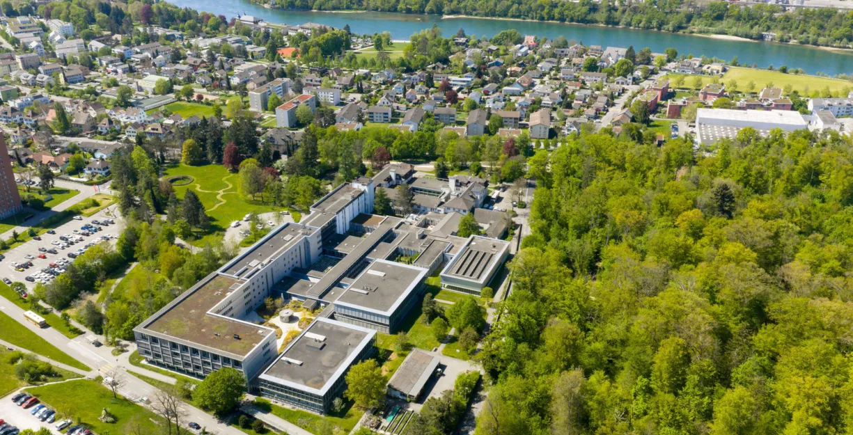 Luftaufnahme eines grünen Klinik- bzw. Universitätsgeländes mit mehreren Gebäudekomplexen, Innenhöfen und Zufahrtsstraßen, umgeben von Wohngebieten und Flusslauf.