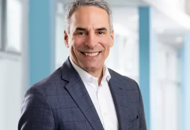 Mann mit grauen Haaren, lächelnd, in einem karierten Anzug und weißem Hemd, vor einem hellen, modernen Hintergrund mit blauen Akzenten.