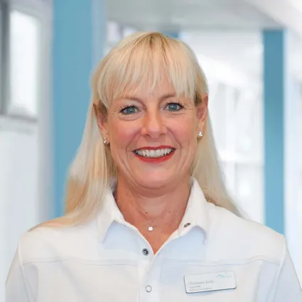 Frau mit blonden Haaren und blauen Augen, lächelnd, trägt ein weißes Oberteil und ein Namensschild. Hintergrund in sanften Blau- und Grautönen.