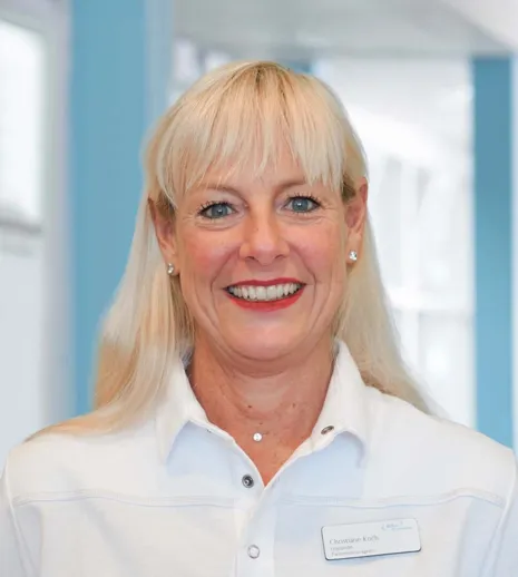 Frau mit blonden Haaren und blauen Augen, lächelnd, trägt ein weißes Oberteil und ein Namensschild. Hintergrund in sanften Blau- und Grautönen.