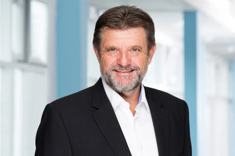 Mann mit Bart und lächelndem Gesicht, trägt einen schwarzen Anzug und ein weißes Hemd, vor einem hellen, modernen Hintergrund.