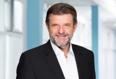 Mann mit Bart und lächelndem Gesicht, trägt einen schwarzen Anzug und ein weißes Hemd, vor einem hellen, modernen Hintergrund.