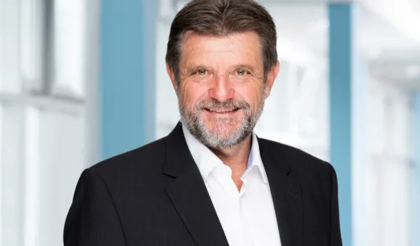 Mann mit Bart und lächelndem Gesicht, trägt einen schwarzen Anzug und ein weißes Hemd, vor einem hellen, modernen Hintergrund.