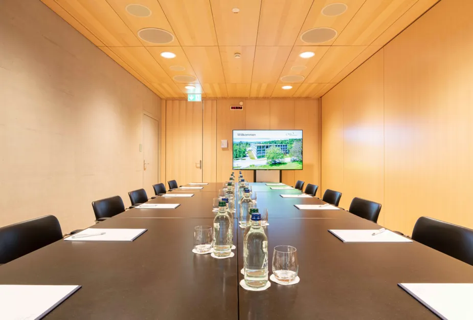 Konferenzraum mit einem langen Tisch, umgeben von schwarzen Stühlen. Auf dem Tisch liegen Notizblätter und Stifte. Im Hintergrund ein Bildschirm mit der Aufschrift „Willkommen“ und einer Abbildung eines Gebäudes. Wassertropfen in Glasflaschen und Gläse...