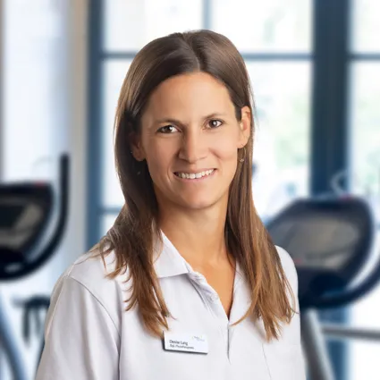 Lächelnde Frau im weißen Polohemd mit Namensschild, im Fitnessstudio vor Trainingsgeräten und großen Fenstern im Hintergrund.