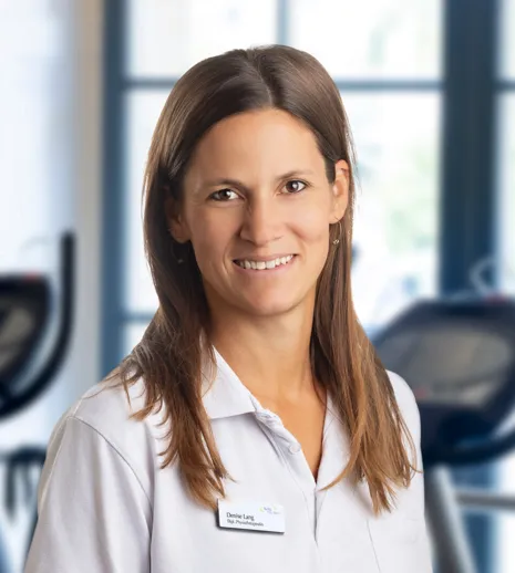 Lächelnde Frau im weißen Polohemd mit Namensschild, im Fitnessstudio vor Trainingsgeräten und großen Fenstern im Hintergrund.
