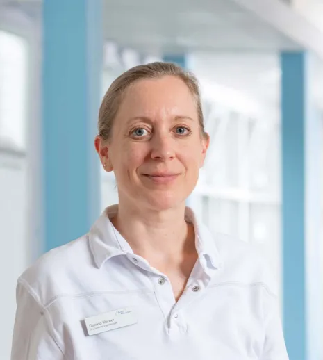Porträt einer Frau im weißen Hemd in einem Büro mit blauen Elementen, freundlich in die Kamera lächelnd.