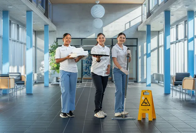 Drei Reinigungskräfte in einer hellen Empfangshalle, mit Handtüchern, Tablett und Wischmopp; gelbes Warnschild „Rutschgefahr“ im Vordergrund.