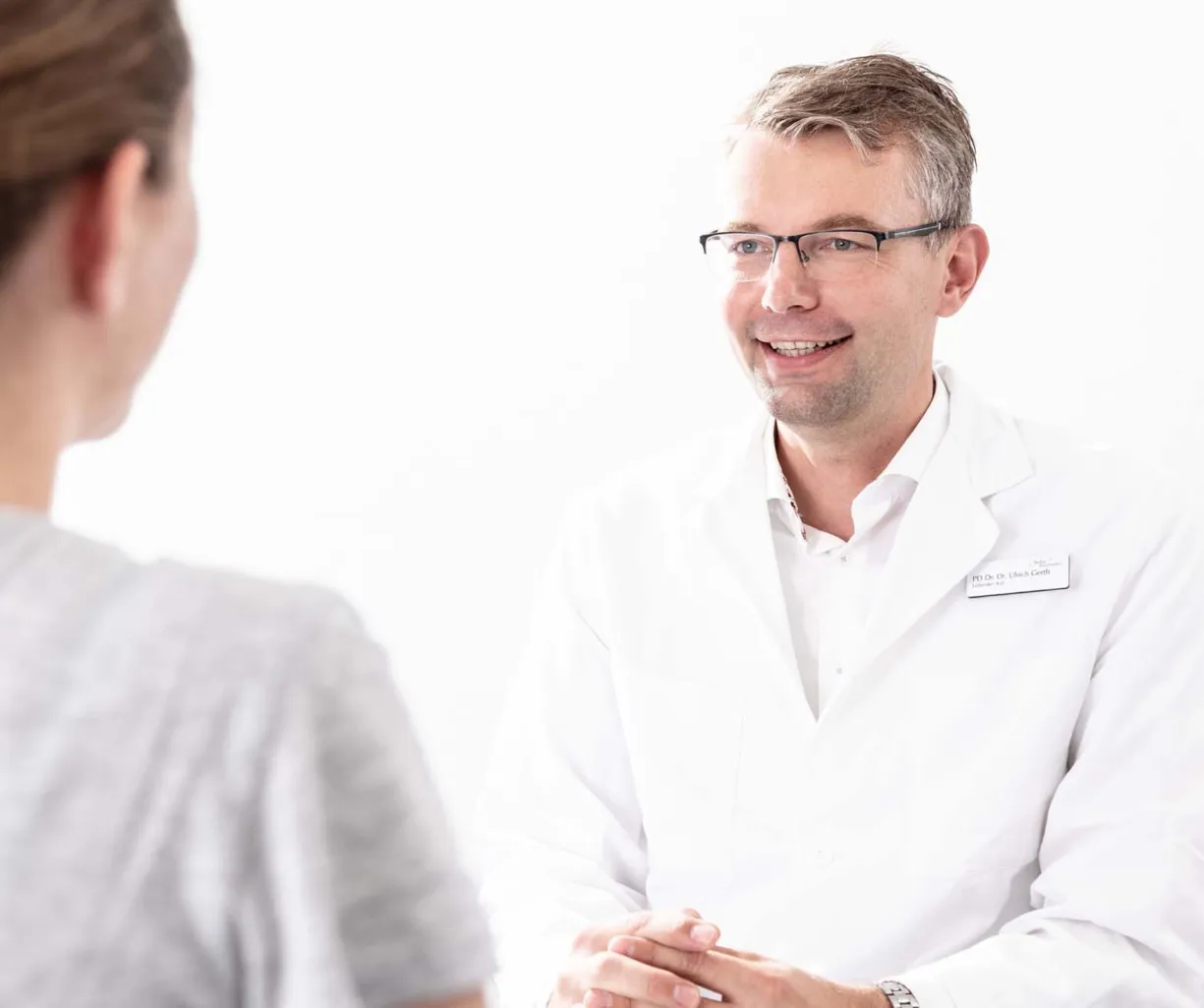 Arzt in weißem Kittel lächelt während eines Gesprächs mit einer Patientin, die ihm den Rücken zuwendet. Helle, neutrale Umgebung.