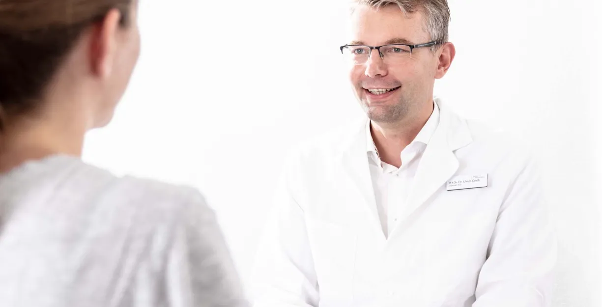 Arzt in weißem Kittel lächelt während eines Gesprächs mit einer Patientin, die ihm den Rücken zuwendet. Helle, neutrale Umgebung.