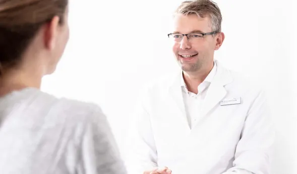 Arzt in weißem Kittel lächelt während eines Gesprächs mit einer Patientin, die ihm den Rücken zuwendet. Helle, neutrale Umgebung.