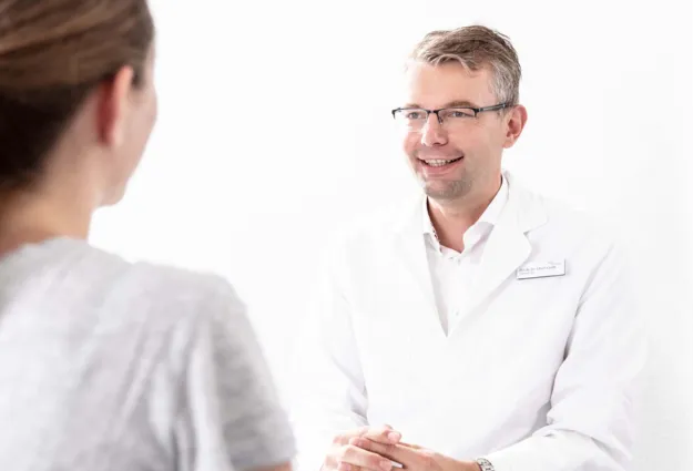 Arzt in weißem Kittel lächelt während eines Gesprächs mit einer Patientin, die ihm den Rücken zuwendet. Helle, neutrale Umgebung.