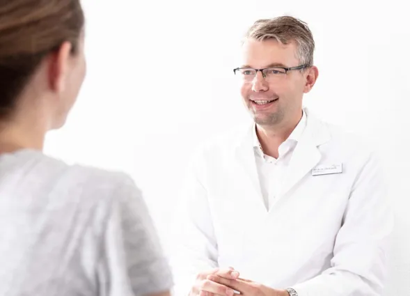 Arzt in weißem Kittel lächelt während eines Gesprächs mit einer Patientin, die ihm den Rücken zuwendet. Helle, neutrale Umgebung.