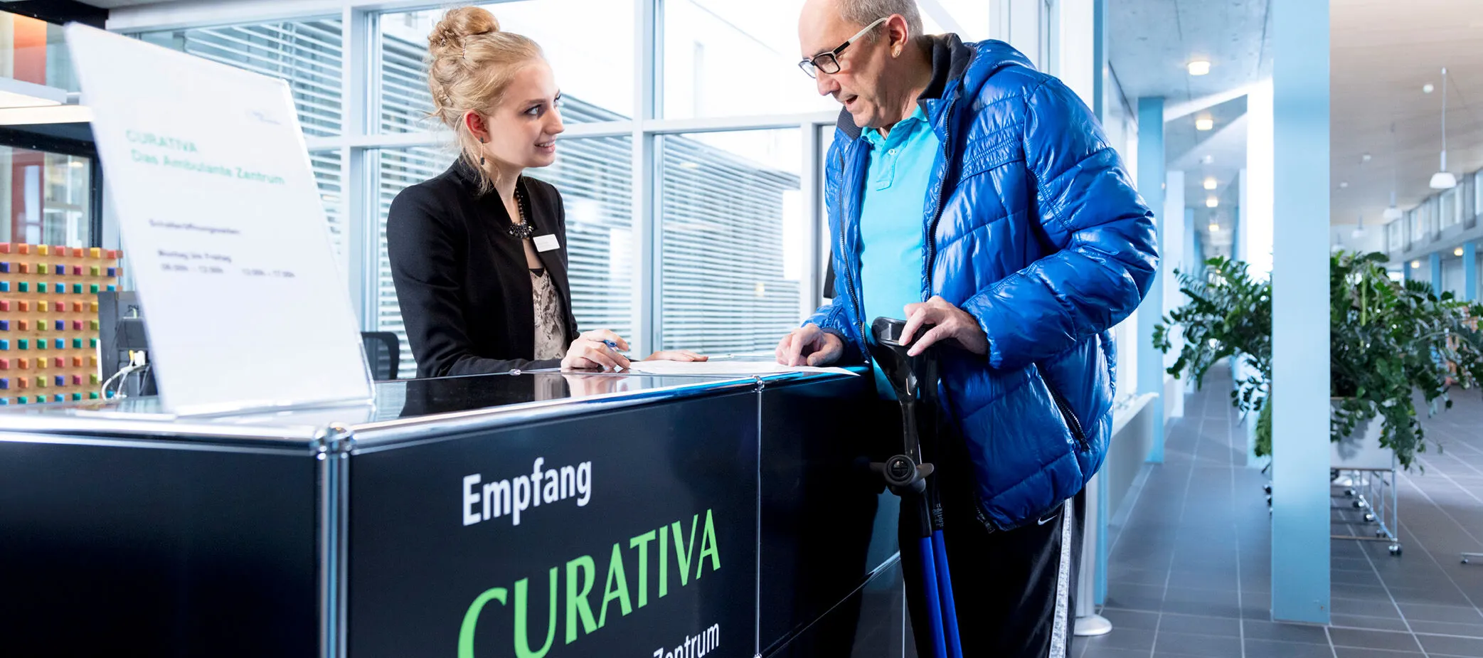 Empfangsbereich eines ambulanten Zentrums mit einer jungen Frau in schwarzer Jacke, die einem älteren Mann in blauer Jacke und mit Gehstock Informationen gibt. Im Hintergrund sind große Fenster und Pflanzen sichtbar.