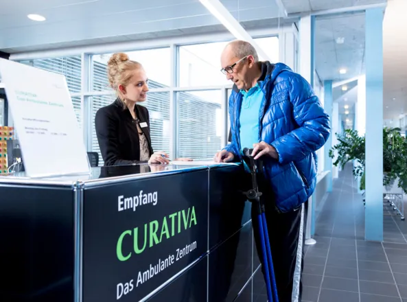 Empfangsbereich eines ambulanten Zentrums mit einer jungen Frau in schwarzer Jacke, die einem älteren Mann in blauer Jacke und mit Gehstock Informationen gibt. Im Hintergrund sind große Fenster und Pflanzen sichtbar.