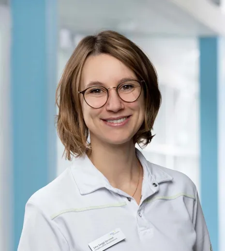 Frau mit kurzen, braunen Haaren und Brille, trägt ein weißes Oberteil mit einem Namensschild, lächelt vor einem hellen, modernen Hintergrund.