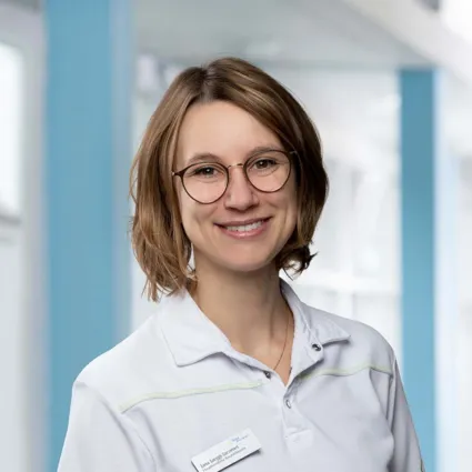 Frau mit kurzen, braunen Haaren und Brille, trägt ein weißes Oberteil mit einem Namensschild, lächelt vor einem hellen, modernen Hintergrund.