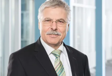 Mann mit grauen Haaren und Brille, trägt einen schwarzen Anzug und eine gestreifte Krawatte, lächelt vor einem modernen, hellen Hintergrund mit großen Fenstern.