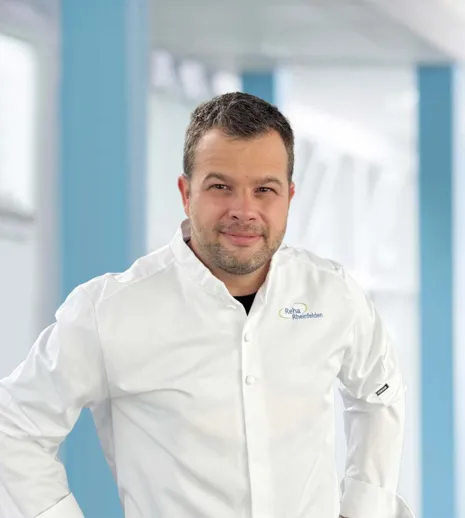 Mann in weißem Kochjackett mit Logo, lächelt und steht in einem hellen, modernen Raum mit blauen Akzenten.