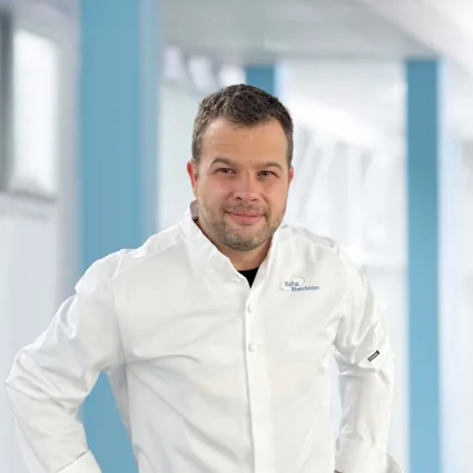 Mann in weißem Kochjackett mit Logo, lächelt und steht in einem hellen, modernen Raum mit blauen Akzenten.