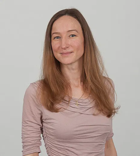 Porträt einer lächelnden Frau mit langen braunen Haaren und rosafarbenem Oberteil vor grauem Hintergrund.