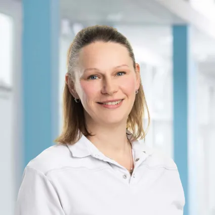 Lächelnde Frau mit langen, hellen Haaren, trägt ein weißes Oberteil, vor einem hellen, modernen Hintergrund mit blauen Akzenten.