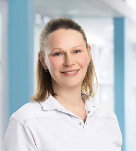 Lächelnde Frau mit langen, hellen Haaren, trägt ein weißes Oberteil, vor einem hellen, modernen Hintergrund mit blauen Akzenten.