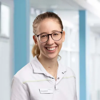 Lächelnde Person mit Brille in einem weißen Oberteil, im Hintergrund eine helle, moderne Umgebung mit blauen Akzenten.