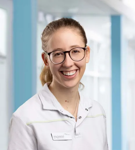 Lächelnde Person mit Brille in einem weißen Oberteil, im Hintergrund eine helle, moderne Umgebung mit blauen Akzenten.