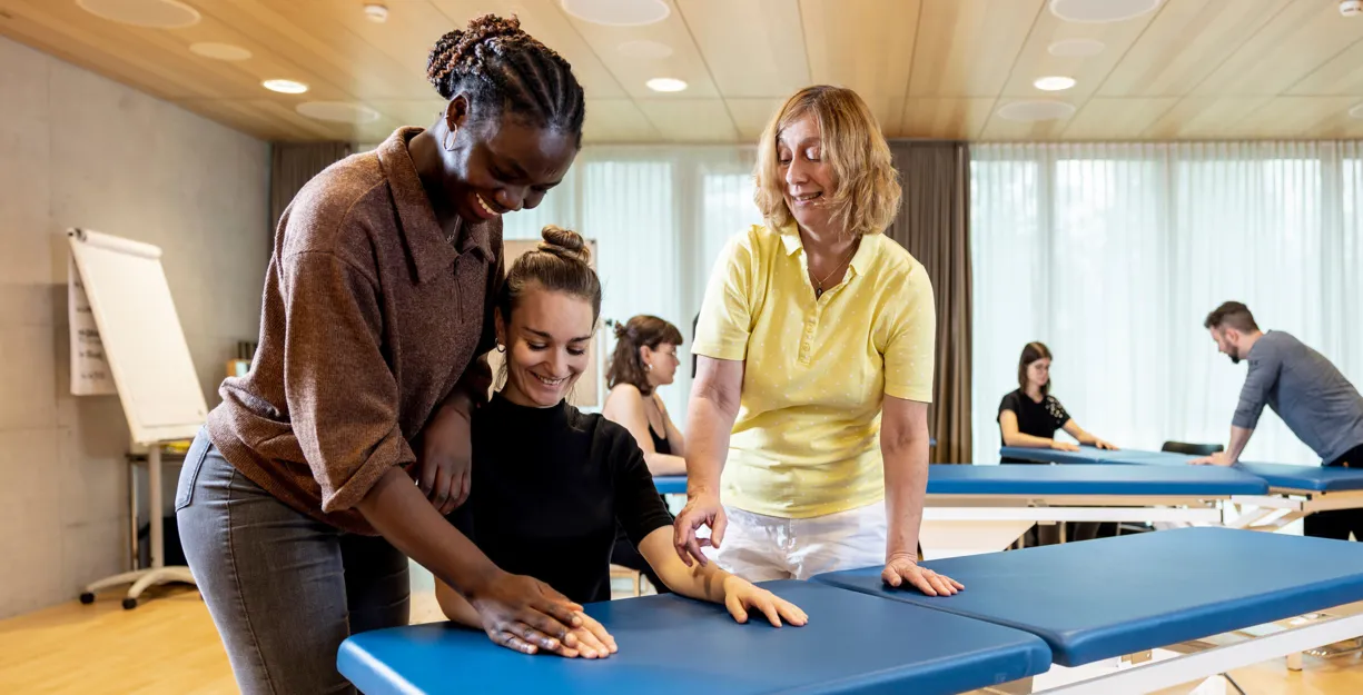 Drei Personen in einem Schulungsraum, zwei Frauen helfen einer dritten Frau, die auf einer blauen Liege sitzt. Im Hintergrund sind weitere Personen an ähnlichen Liegen zu sehen. Der Raum hat große Fenster mit Tageslicht und eine moderne Einrichtung.