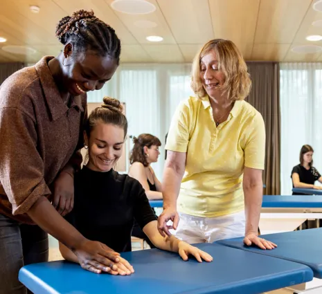 Drei Personen in einem Schulungsraum, zwei Frauen helfen einer dritten Frau, die auf einer blauen Liege sitzt. Im Hintergrund sind weitere Personen an ähnlichen Liegen zu sehen. Der Raum hat große Fenster mit Tageslicht und eine moderne Einrichtung.