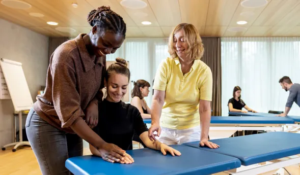 Drei Personen in einem Schulungsraum, zwei Frauen helfen einer dritten Frau, die auf einer blauen Liege sitzt. Im Hintergrund sind weitere Personen an ähnlichen Liegen zu sehen. Der Raum hat große Fenster mit Tageslicht und eine moderne Einrichtung.