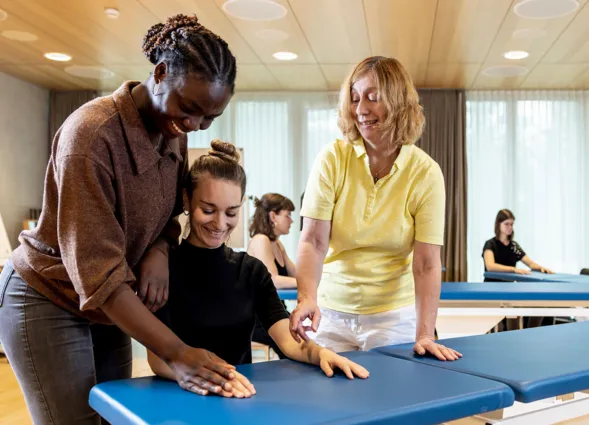 Drei Personen in einem Schulungsraum, zwei Frauen helfen einer dritten Frau, die auf einer blauen Liege sitzt. Im Hintergrund sind weitere Personen an ähnlichen Liegen zu sehen. Der Raum hat große Fenster mit Tageslicht und eine moderne Einrichtung.