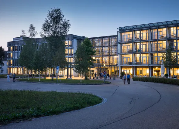 Modernes Gebäudeensemble mit verglasten Fassaden und warm beleuchteten Innenräumen bei Dämmerung, umgeben von Bäumen, Wegen und mehreren Passanten.